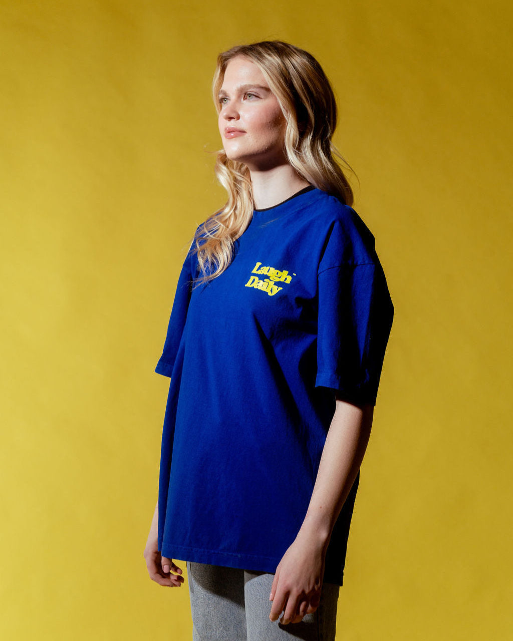 LAUGH DAILY ICON TEE - COBALT BLUE worn by a tall blonde female model posing with a yellow backdrop