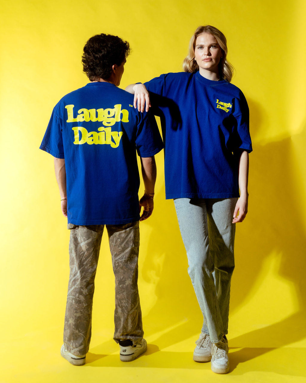 LAUGH DAILY ICON TEE - COBALT BLUE worn by two models male and female standing together with a yellow backdrop posing in shirts